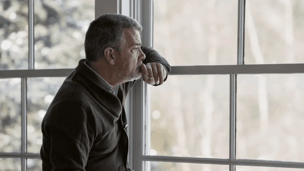 Elderly man looking out a window during the winter, appearing thoughtful and melancholic.