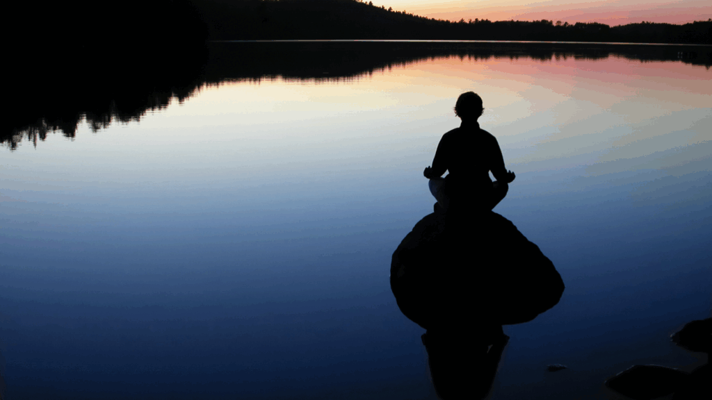 Person meditating by a calm lake at sunset, practicing intentional awareness to rewire fear responses using neuroplastic momentum.
