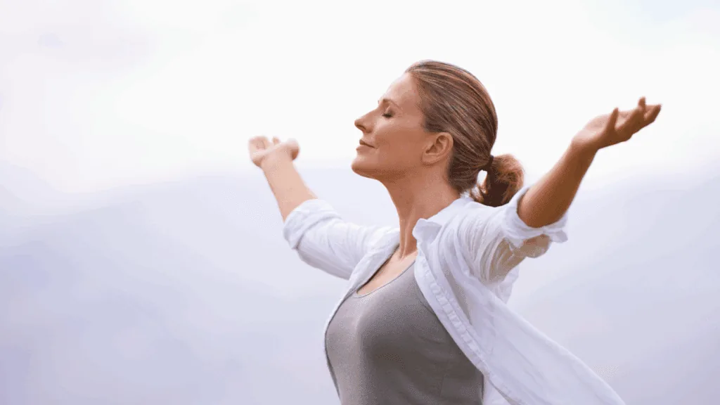 A woman standing outdoors with arms raised, representing steadier energy and motivation.