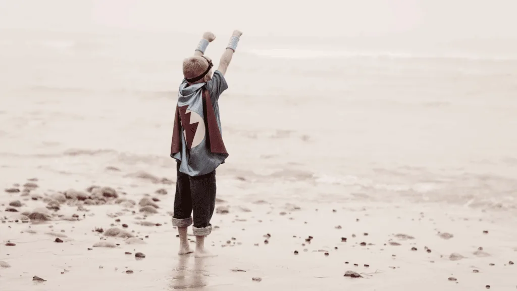 Child in a superhero cape standing on a beach, symbolizing confidence and the roots of presence.