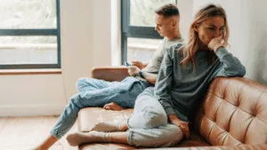 Couple sitting apart on a couch looking distant, showing the emotional impact of what if thinking on relationships.
