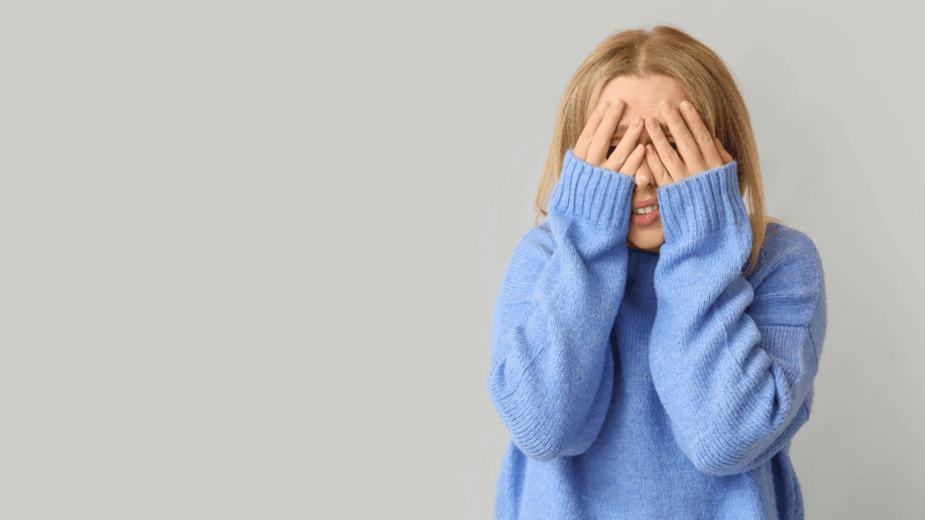 Woman in a blue sweater covering her face, expressing insecurity about feeling like a loser.