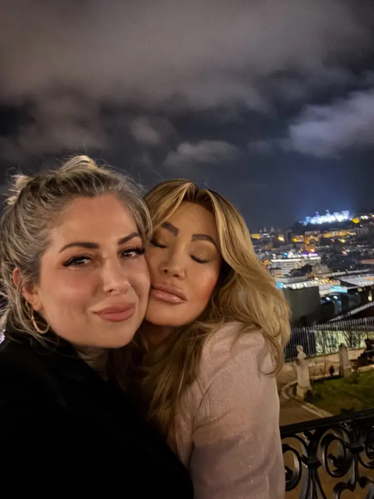 Dr. Sydney Ceruto embracing close friend Mila at night in Lisbon with city lights showing deep authentic friendship