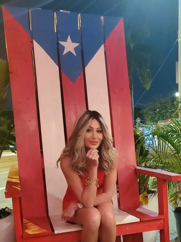 Dr. Sydney Ceruto smiling in a red dress seated on an oversized Puerto Rican flag chair during a night out in Puerto Rico