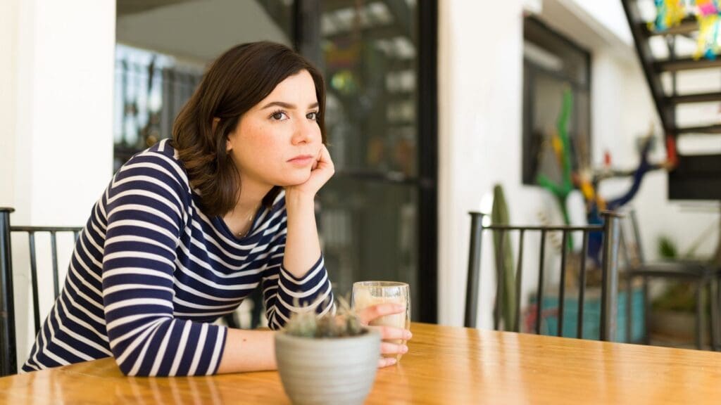 Woman in a striped shirt sits alone at a table with a drink, resting her head on her hand, expressing the isolation and frustration behind the keyword Meet Anyone.