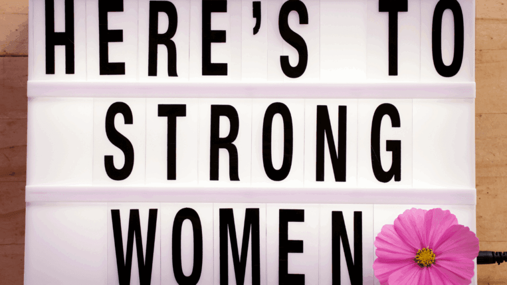Balancing Strength and Empowerment in Women Woman holding a sign saying "Here's to strong women" symbolizing the balance of strength and empowerment in women.