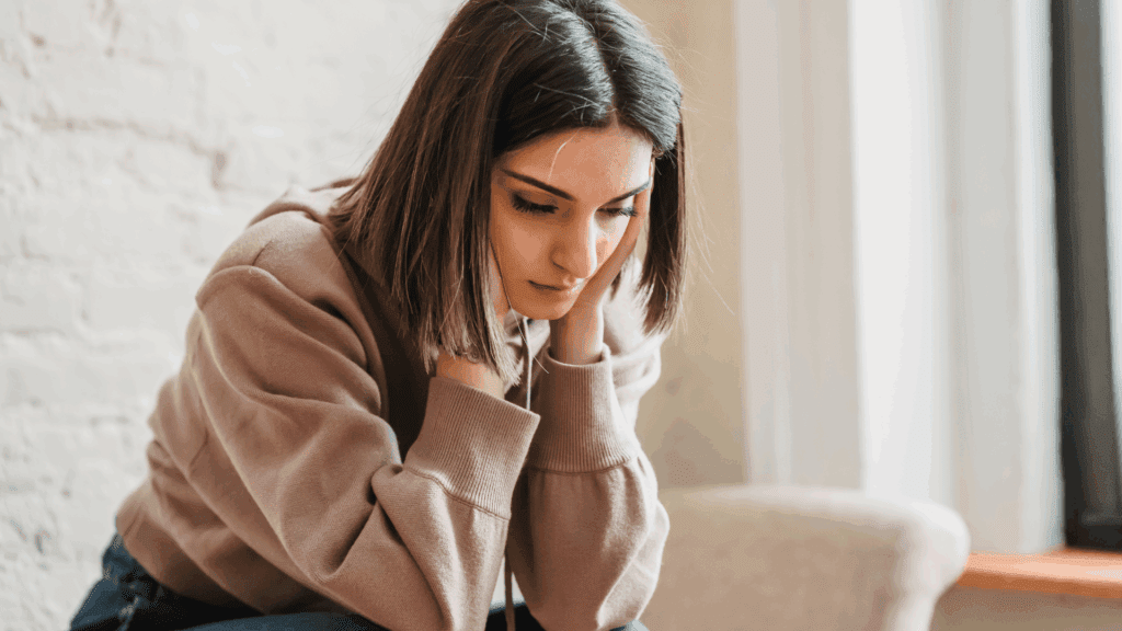 Young woman sitting indoors with her head in her hands, appearing deep in thought and feeling empty.