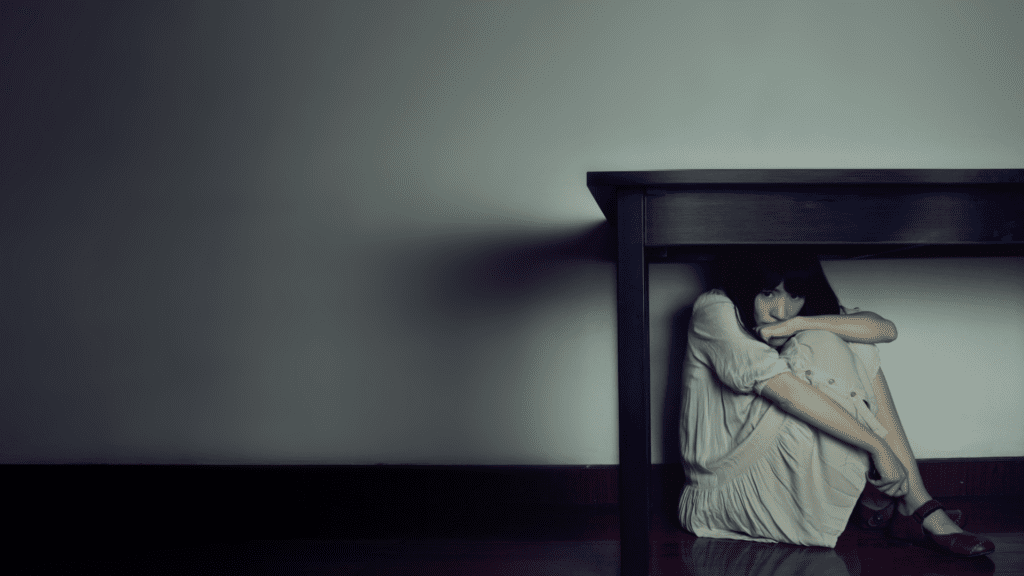  A woman sitting under a table in a dimly lit room, symbolizing the isolating effects of fear and anxiety.


