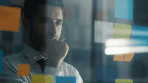 Thoughtful businessman looking at sticky notes on glass wall, reflecting on goals and strategies while feeling stuck.