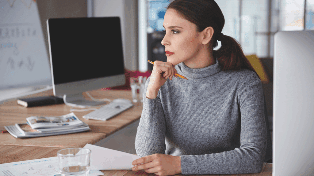 Professional woman in grey sweater thoughtfully analyzing work at desk representing neuroscience based mindset coaching client reflection
