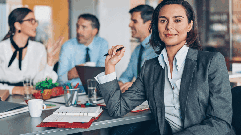 A confident executive woman sits at a meeting table with colleagues in the background, embodying success from neuroscience coaching for executives.

