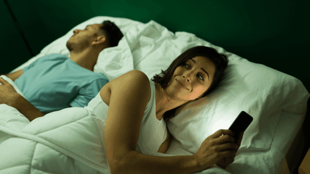 Woman in bed looking at smartphone while man sleeps beside her, illustrating emotional disconnection and digital temptation in dopamine and infidelity within relationships.

