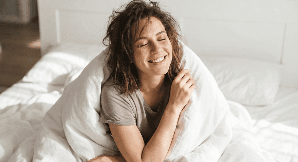 Smiling young woman sitting up in bed wrapped in white bedding, eyes closed, conveying contentment after self-pleasure.