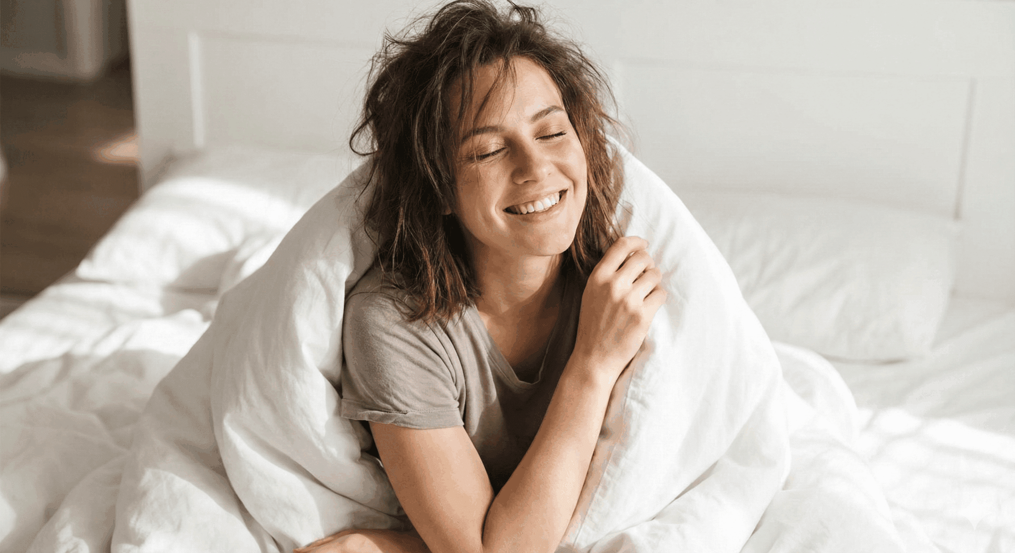 Smiling young woman sitting up in bed wrapped in white bedding, eyes closed, conveying contentment after self-pleasure.