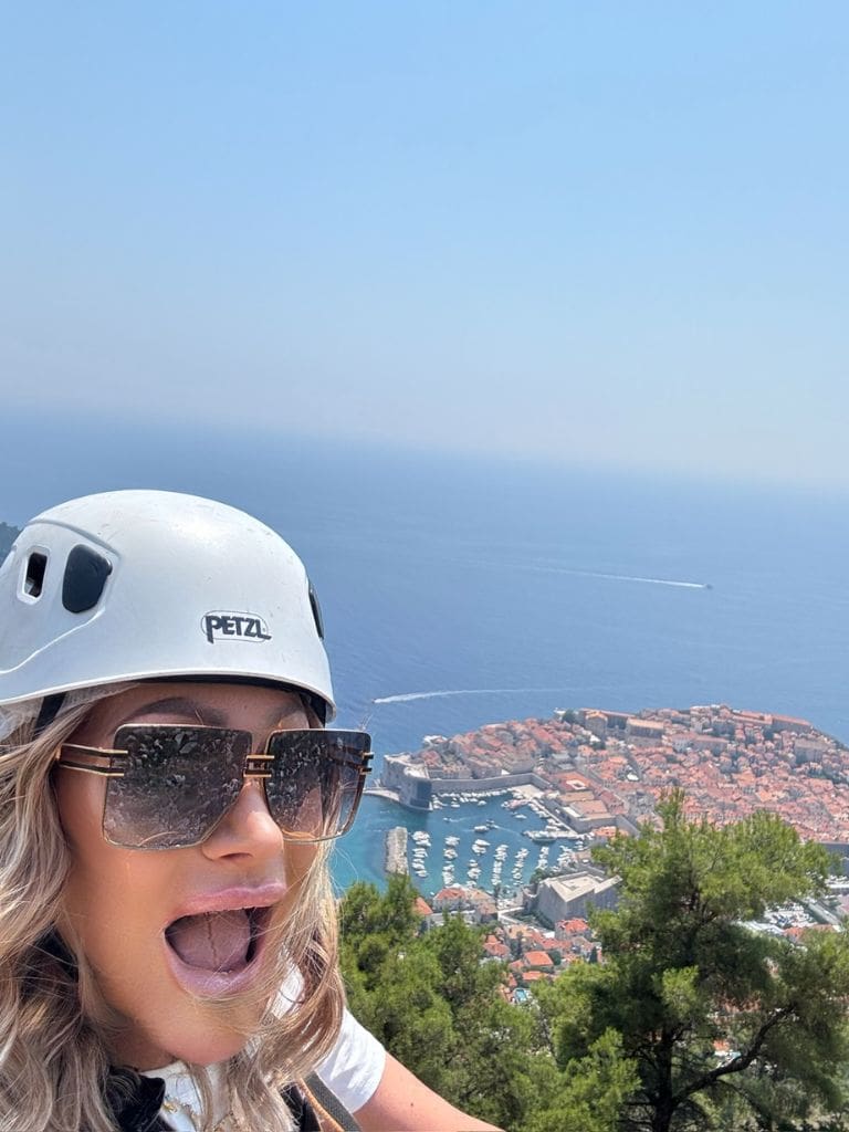 Dr. Sydney Ceruto wearing a safety helmet taking an excited selfie high above the old town and harbor of Dubrovnik, Croatia during a zip-line adventure